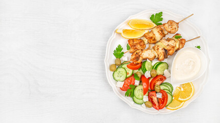 Chicken skewers served with fresh vegetable salad, lemon slices, and creamy sauce on a white plate over a bright wooden background. top view. copy space