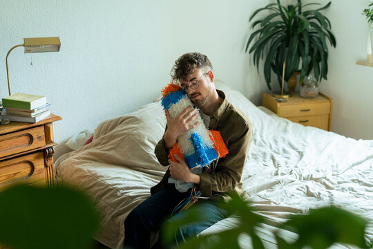 Young man with eyes closed hugging rocketship shaped pinata sitting on bed at home