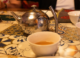 Traditional metal teapot with tea cup. Cozy table with colorful tiles. Warm ambient lighting in restaurant. Tea time setting close-up. Relaxing beverage moment indoors.
