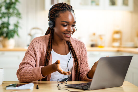 Online Tutoring. Smiling black female tutor in headset using laptop in kitchen, young african american teacher having video call with students, talking and gesturing at web camera, free space