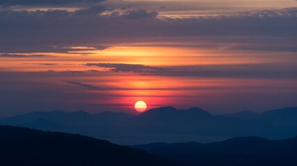 Fototapeta premium Sun setting behind layered blue mountains with orange sky
