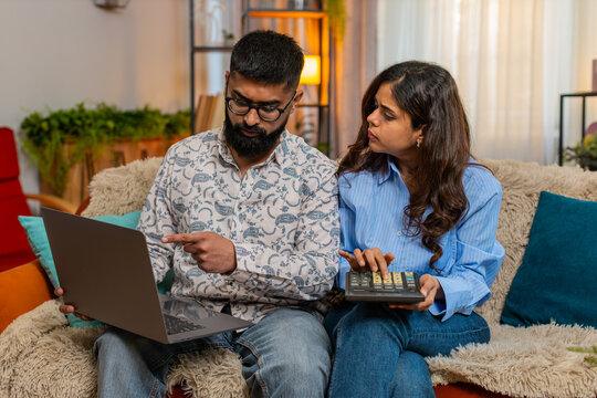 Indian couple man and woman sitting on home sofa with laptop and calculator calmly discussing expenses and future savings. Young family working on home budget plan with focus and understanding indoors