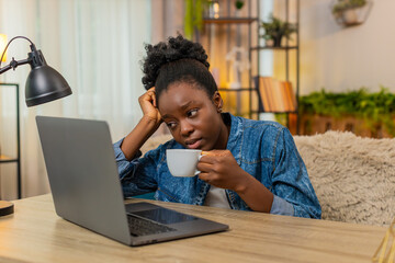 African American sleepy woman sits at table on home sofa drinks coffee tiredly after sleepless night trying to energize herself. Black girl prepares to start remote workday using laptop stay focused