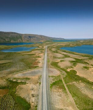 Aerial view of a stark road cuts through a vibrant landscape of green and brown fields, leading towards serene lakes under a clear blue sky, Raufarh&Atilde;&para;fn, Nor&Atilde;&deg;ur&Atilde;&frac34;ing, Iceland.
