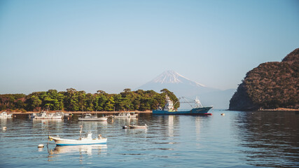 Heda, Japan - 01 January 2025: View of a serene bay dotted with fishing boats, the majestic Mount Fuji rising distantly behind lush, verdant trees and calm, blue waters.