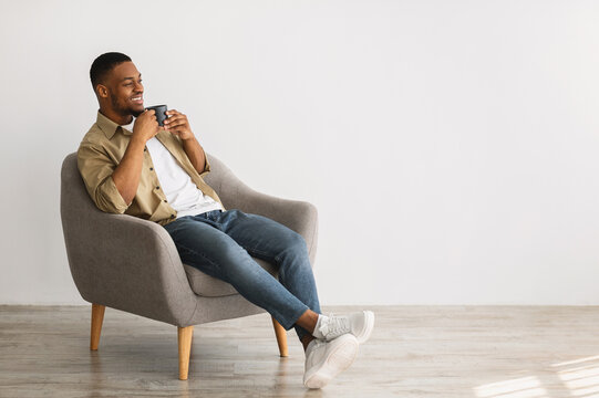 Relaxed Black Guy Enjoying Morning Coffee Sitting In Gray Chair Looking Aside At Copy Space Indoors. Comfort Of Minimalism Lifestyle. Weekend Leisure Concept. Advertisement Banner