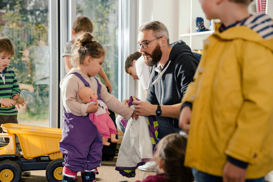 Pre-school teacher helping girl putting on rainwear in kindergarten