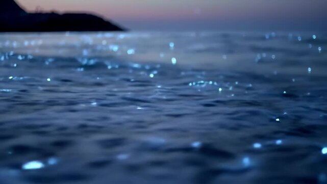 Magical close-up of glowing blue bioluminescent plankton in gentle ocean waves on a sandy beach at dusk