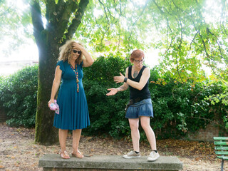 Mature women conversing on park wall, one shading eyes and laughing, other animatedly gesturing during storytelling moment