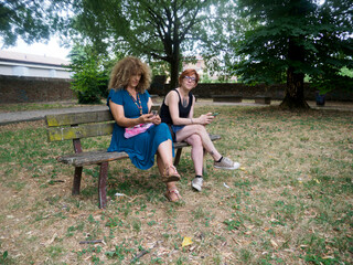 Fototapeta premium Two women sitting on a park bench outdoors using their mobile phones with trees and grass in the background on a sunny day enjoying leisure time