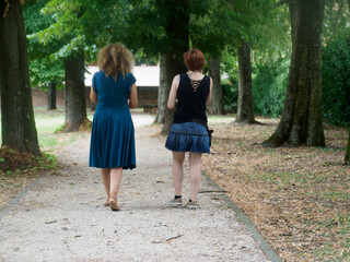 Friends walking together along tree lined park path, sharing intimate conversation amid verdant...