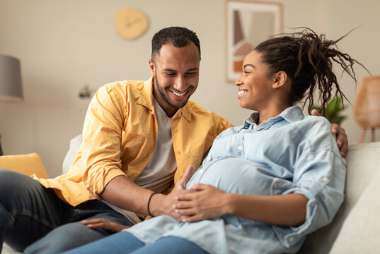 Happy african american couple expecting baby, pregnant black spouses embracing on couch at home. Smiling husband and wife resting in living room - Powered by Adobe