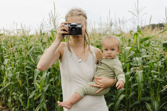 Mother photographing through camera carrying toddler girl in front of crops