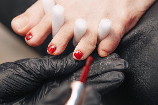 Red nail polish on toes. Pedicure procedure in salon. Beautician in black gloves applying polish. Female feet with separators. Closeup of foot care service. Beauty and hygiene treatment indoors.