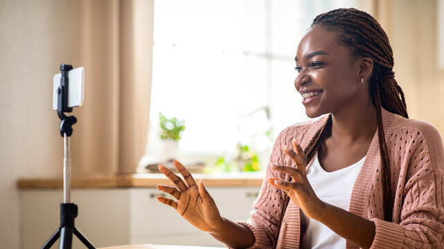 Young african american female blogger recording new video vlog on smartphone at home, using tripod, beautiful smiling black lady making live stream, talking and gesturing at camera, copy space - Powered by Adobe
