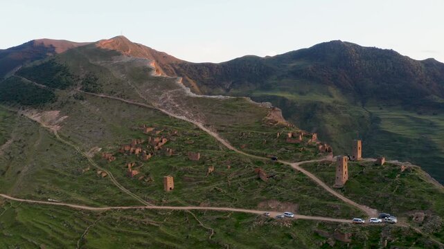 Aerial view of ancient stone towers dotting the green hillsides, contrasting with the winding roads and parked cars, Goor, Republic of Dagestan, Russia.