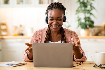 Teleconference. Positive black woman in headset having video call on laptop in kitchen, talking and gesturing at computer web camera, enjoying online conversation and distant communication, free space