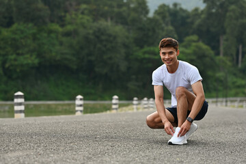 A Smiling athlete crouching down to tie his running shoes on a road, preparing for an outdoor...