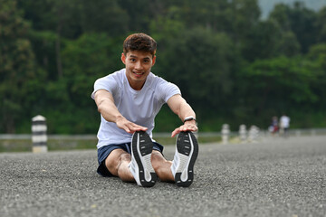 A Fit man sitting on the road performing a forward stretch, preparing for an outdoor workout in a...