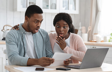 Shocked black couple in kitchen stressed with financial problems, doing calculations of family budget together. Frustrated african american spouses sitting at table looking at loan documents