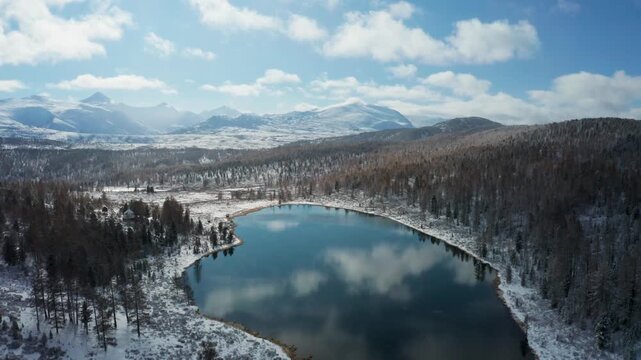 Aerial view of a pristine lake reflecting the sky, surrounded by snow-dusted trees and distant mountains under a blanket of white clouds, Ulagan, Altai Republic, Russia.