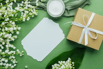 Blank label card, candle, and lily of the valley flowers with gift box on green background