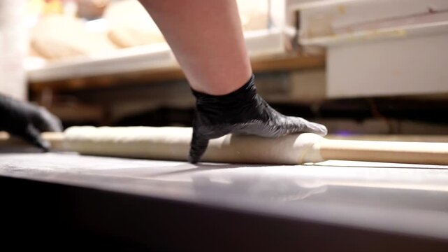 A chef rolls dough with a rolling pin on a floured metal surface, wearing black gloves for hygiene.