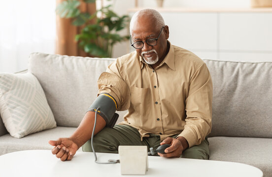Hypertension Problem In Senior Age. African American Man Measuring Arterial Blood Pressure Using Sphygmomanometer Cuff Sitting On Couch At Home. Healthcare, Health Issue