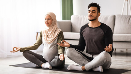 Pregnant muslim couple practicing prenatal yoga at home, meditating together with closed eyes,...