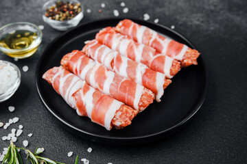 Raw minced meat rolls wrapped in bacon on black plate with spices