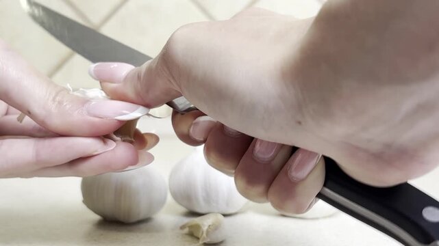 Close-up of woman's hand peeling garlic with a knife on kitchen surface, food preparation process, cooking at home, fresh ingredient, natural light, healthy eating, rustic kitchen scene