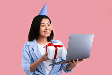 Cheerful young asian woman with party hat holding present and showing box to laptop screen, standing at studio, celebrating or greeting with birthday online during quarantine and lockdown
