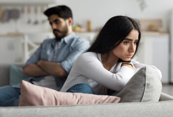Fototapeta premium Negative emotions, problems, quarrel and divorce. domestic scandal during covid-19. Upset offended young arab wife and husband turned away and ignoring each other, silent in living room interior