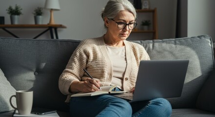 Focused Senior Woman Working on Laptop and Writing in a Notebook on a Couch
