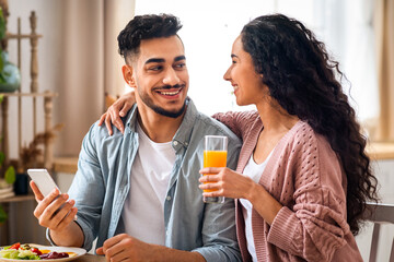 Happy Young Arab Couple Using Smartphone During Breakfast In Kitchen, Cheerful Middle Eastern Lovers Browsing Social Networks On Mobile Phone While Enjoying Tasty Morning Meal At Home, Closeup Shot