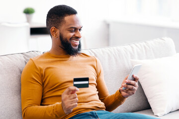 African American Guy Using Credit Card And Smartphone Making Transaction Or Payment Online Via Mobile Banking Application Sitting On Couch At Home. Shopping App And E-Commerce. Selective Focus