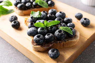 Sweet homemade toasts with chocolate paste, nuts, blueberry and aromatic mint leaves on the serving board on dark stone background. Tasty summer dessert.