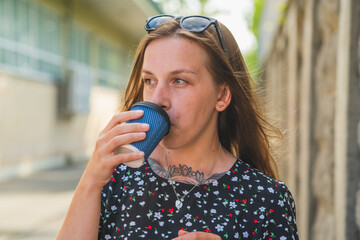 A woman sips coffee from a blue cup while strolling through a sunlit alley She wears a floral dress...