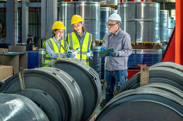 Industrial factory workers wea safety helmets and vests inspecting large metal coils and cylinders...