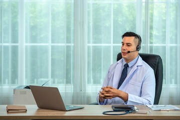 Confident male doctor in white coat and stethoscope around neck wearing headset while sitting at...
