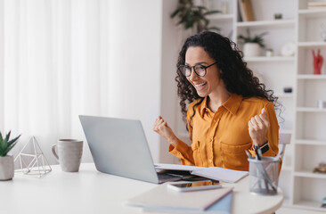 Joyful Middle Eastern Woman Shaking Hands Using Laptop Computer Celebrating Business Success Working Online Sitting At Desk At Home. Remote Job And Freelance, Big Luck Concept. Side View