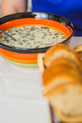 A vibrant bowl of cold okroshka soup filled with herbs and vegetables is placed on a dining table Freshly baked bread slices are presented on the side enhancing the meal