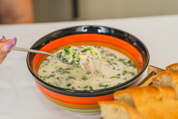 A bowl of cold okroshka soup showcases a blend of herbs vegetables and meat with a spoon lifted to reveal its creamy texture Crusty bread sits nearby enhancing the meal
