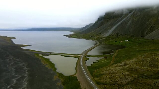 Aerial view of a road cutting through tranquil waters, creating a striking contrast against the black sand beach and the misty mountain, Hvalnes, M&uacute;la&thorn;ing, Iceland.