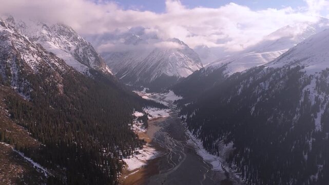Aerial view of snow-capped Boris Yeltsin Peak, with dark green forests and a winding river cutting through the valley, Issyk-Kul Region, Kyrgyzstan.