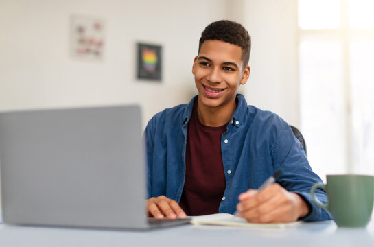 Happy African American teenager guy in blue shirt studying with grey laptop computer, taking notes, enjoying learning at home environment, free space