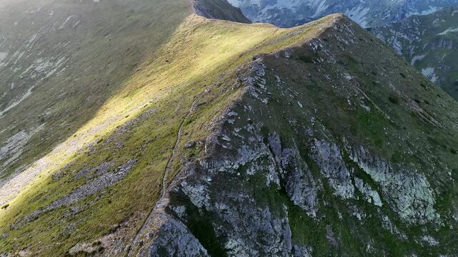 Aerial view of a rugged mountain ridge split by a narrow path, bathed in the warm glow of sunlight contrasting with deep shadows, ziar, zilina Region, Slovakia.