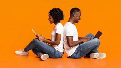 Gadget addiction and relationships concept. Young black family man and woman spending time with gadgets, sitting on floor back to back and using digital tablet and laptop, orange studio background