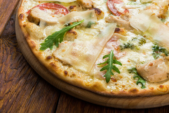 Delicious pizza with chicken, parmesan, tomatoes, white sauce and fresh arugula - thin pastry crust at wooden background, closeup