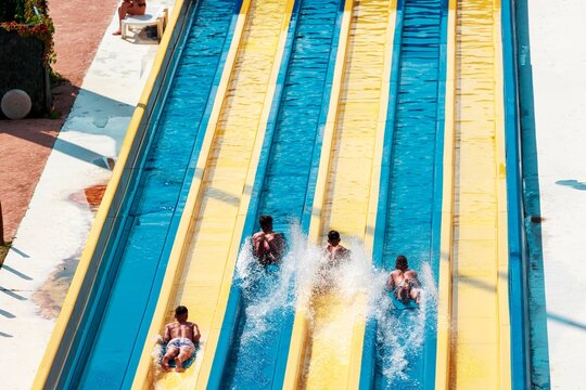 Several people are seen speeding down the bright blue and yellow water slides, generating splashing water as they swiftly descend, enjoying a summer day at the outdoor waterpark in Turkey